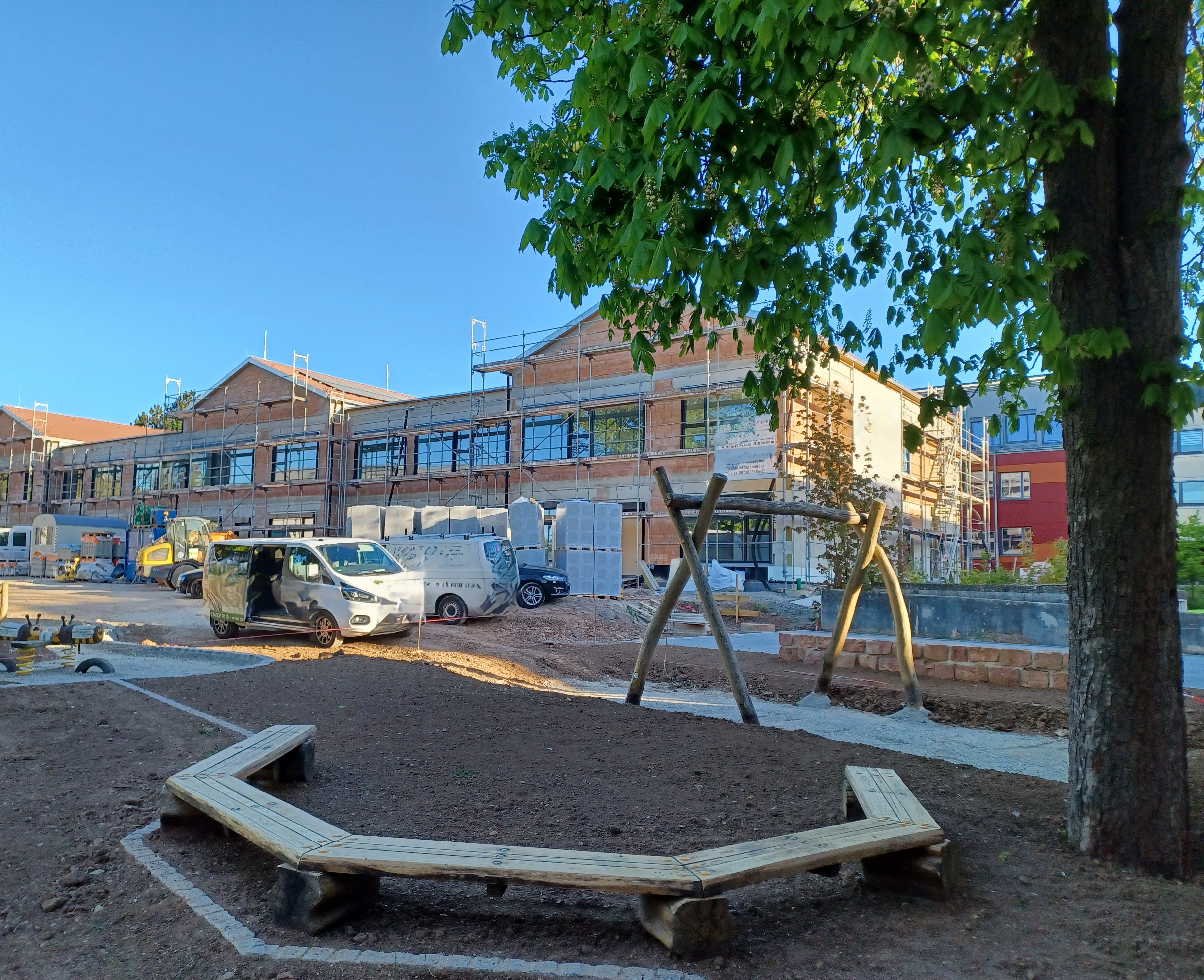 Das Bild zeigt eine Baustelle mit mehreren Gebäuden im Bau. Im Vordergrund befinden sich Baugeräte und Schutt. Im Hintergrund stehen bereits fertige oder fast fertige Häuser mit hellen Fassaden. Ein Baum ist rechts im Bild sichtbar.