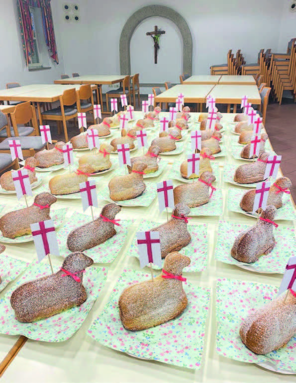 Das Bild zeigt einen Raum mit mehreren Tischen, auf denen längliche Brote in der Form eines Lammes liegen. Jedes Brot hat eine weiße Fahne mit einem roten Kreuz. Im Hintergrund sind Stühle und ein Kreuz an der Wand zu sehen.