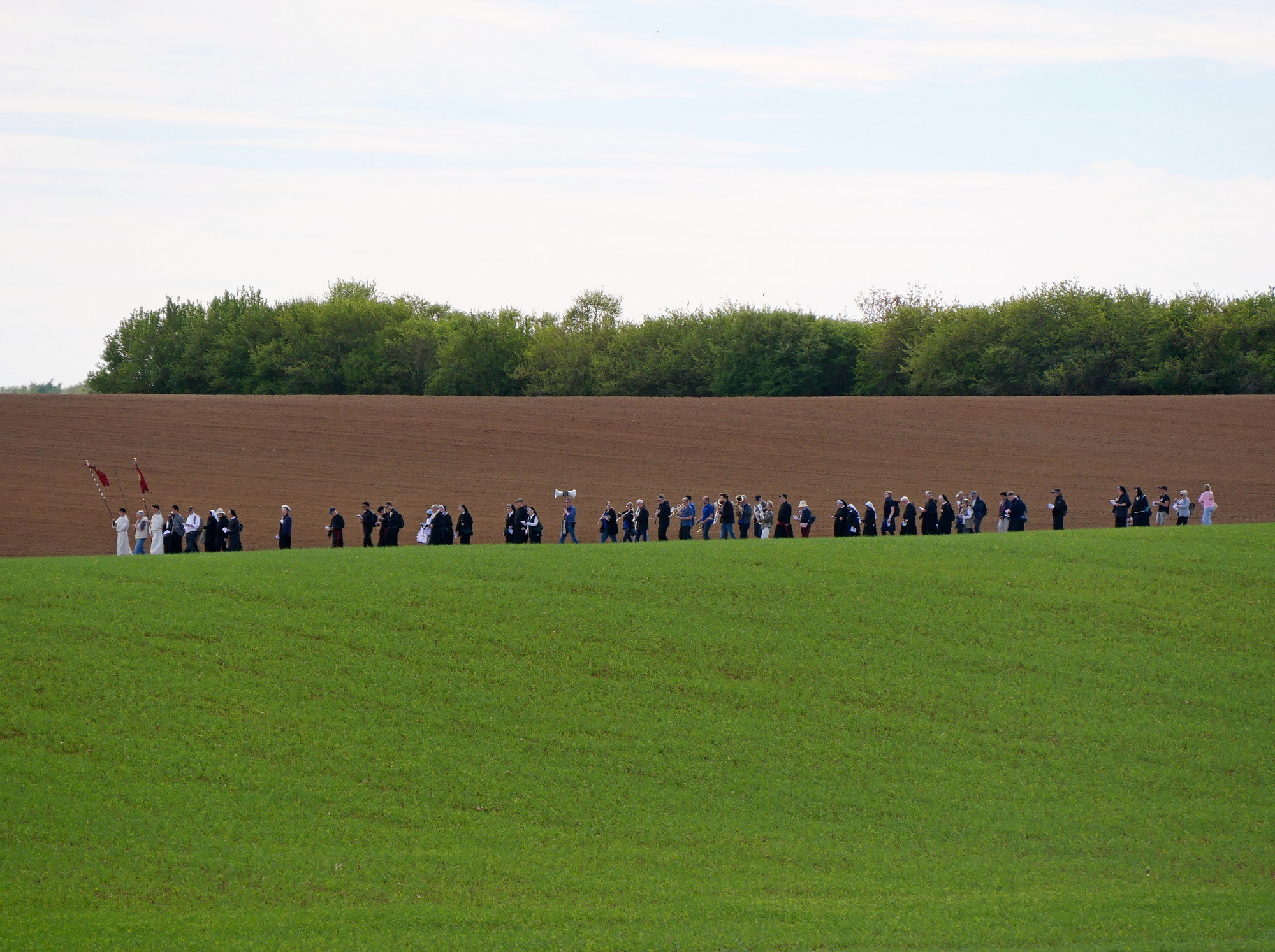 Das Bild zeigt eine Gruppe von Menschen, die sich am Rand eines Feldes versammelt haben. Vor der Wandergruppe ist ein Feld mit grünem Gras, hinter ihnen befindet sich ein frisch gepflügtes Feld und ein Waldstreifen. Einige Personen halten Fahnen oder Schilder. Der Himmel ist klar mit einigen Wolken.