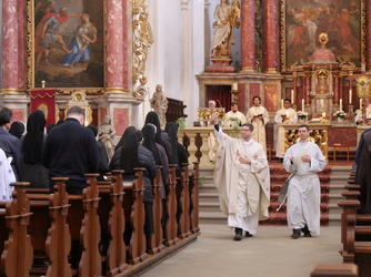 Das Bild zeigt eine katholische Kirche während einer Messe. Im Vordergrund stehen ein Priester und ein Ministrant in weißen Gewändern. Im Hintergrund sind weitere Gläubige, darunter mehrere Nonnen und ein Priester am Altar, zu sehen. Die Kirche ist reich verziert mit Gemälden und Statuen.