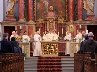 Das Bild zeigt eine feierliche Messe in einer prächtig geschmückten Kirche. Mehrere Priester in weißen Gewändern stehen vor einem reich verzierten Altar. Gläubige sitzen in den Bänken und beten. Der Altar ist mit Kerzen und religiösen Symbolen geschmückt.